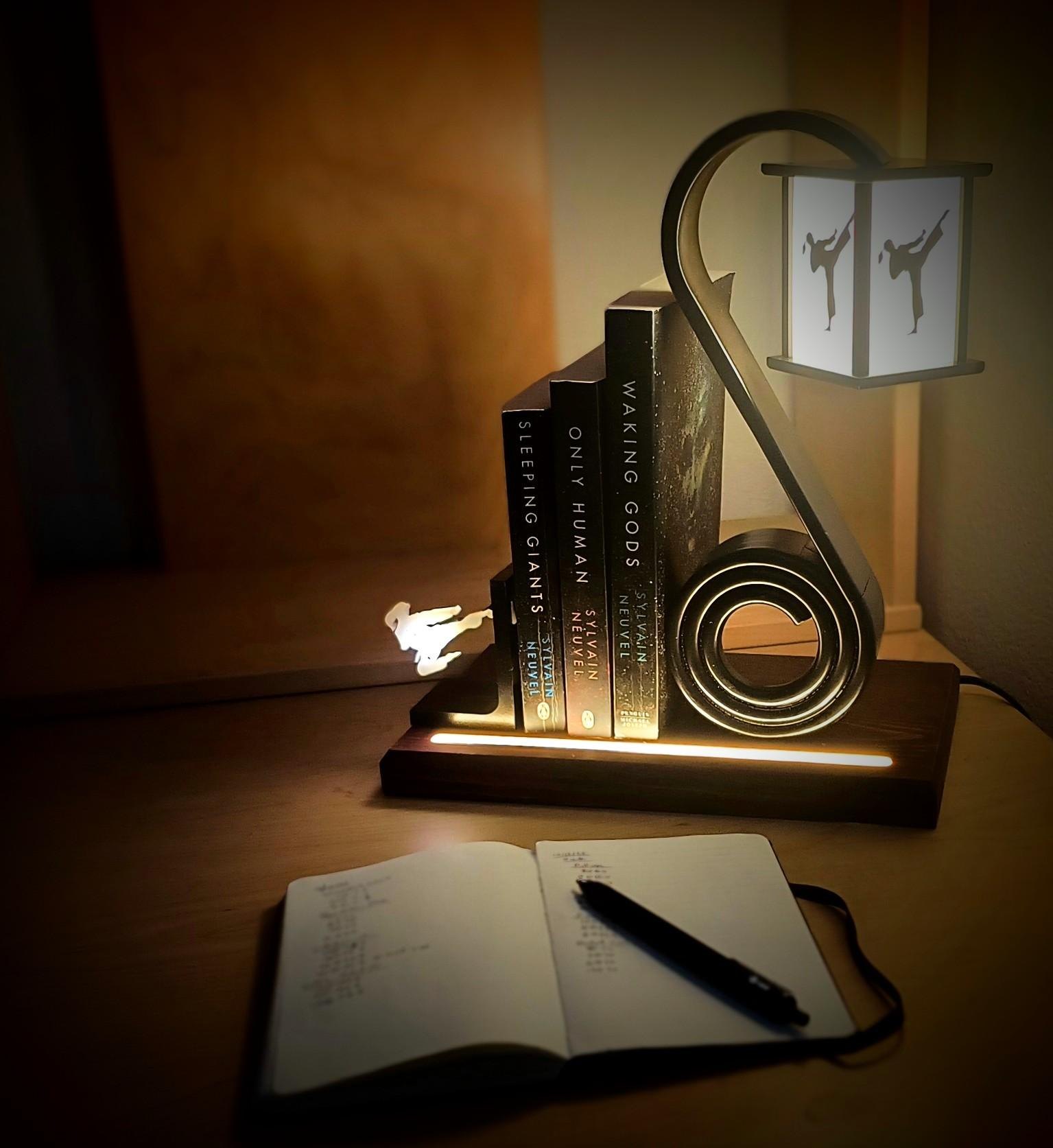The finished lamp lit on a nightstand: a curved black arm rising from a wooden base into a lantern with a kicking figure on its panels, a flat spiral coil at the foot holding a row of books, and a strip of light along the base.