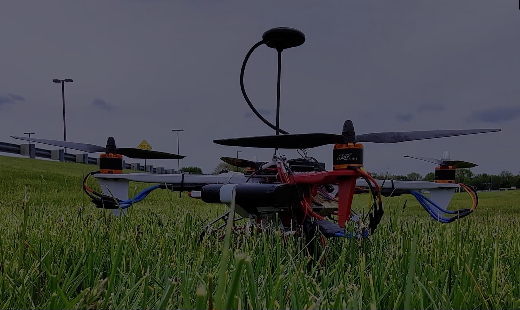 The quadrotor standing in grass at a flying field, carrying its payload mount and antenna, under an overcast sky.