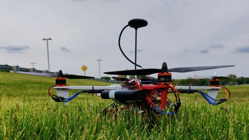 The quadrotor standing in grass at a flying field, its GPS mast upright and payload slung underneath.