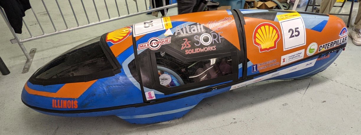 The competition car in a paddock: a low, streamlined orange and blue shell on three wheels, barely knee high, with a clear canopy over the driver's position and sponsor decals along the flank.