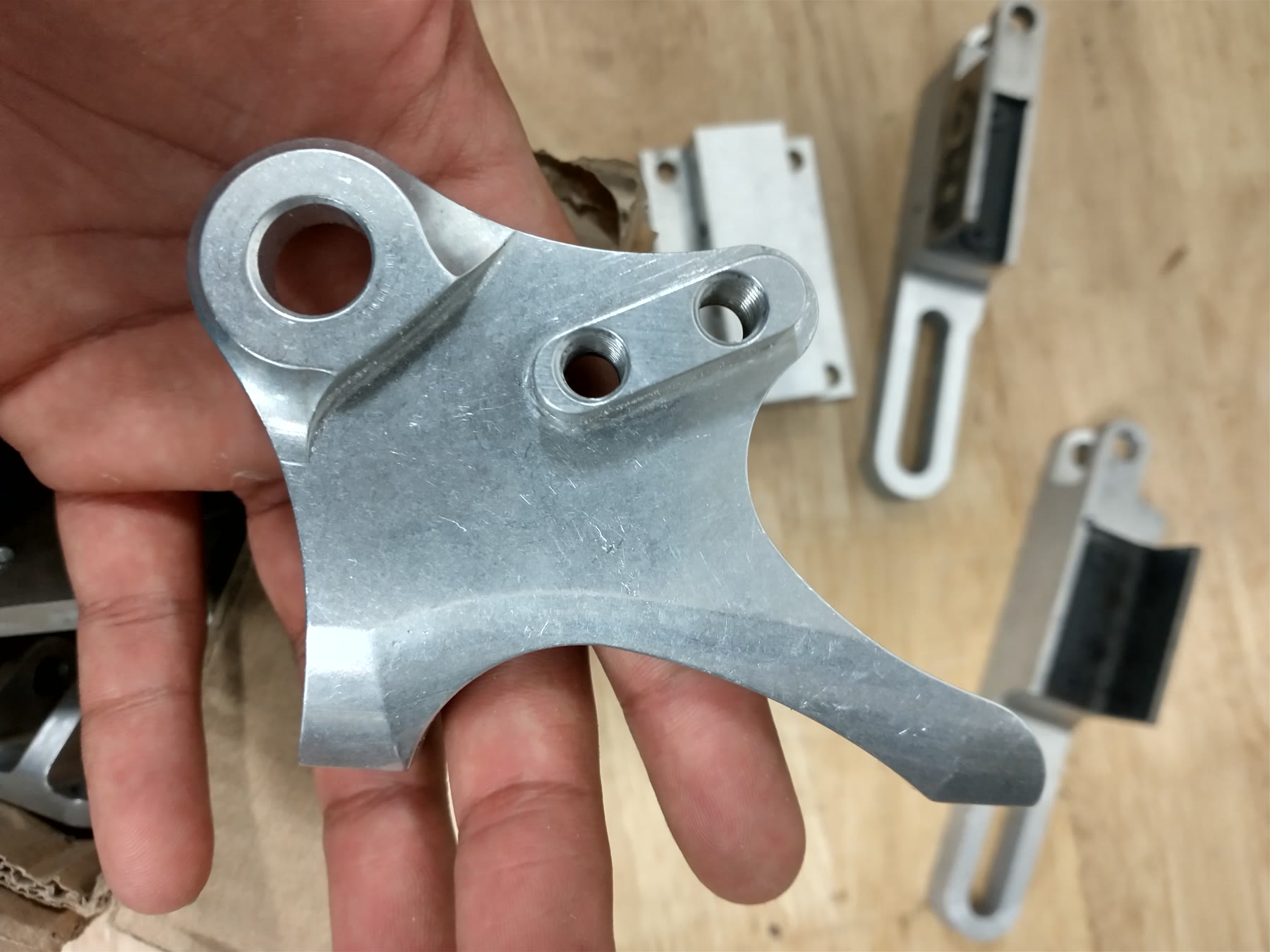 A hand holding a machined aluminium bracket from the rear wheel assembly, with 3D-printed parts and hardware on the bench behind it.